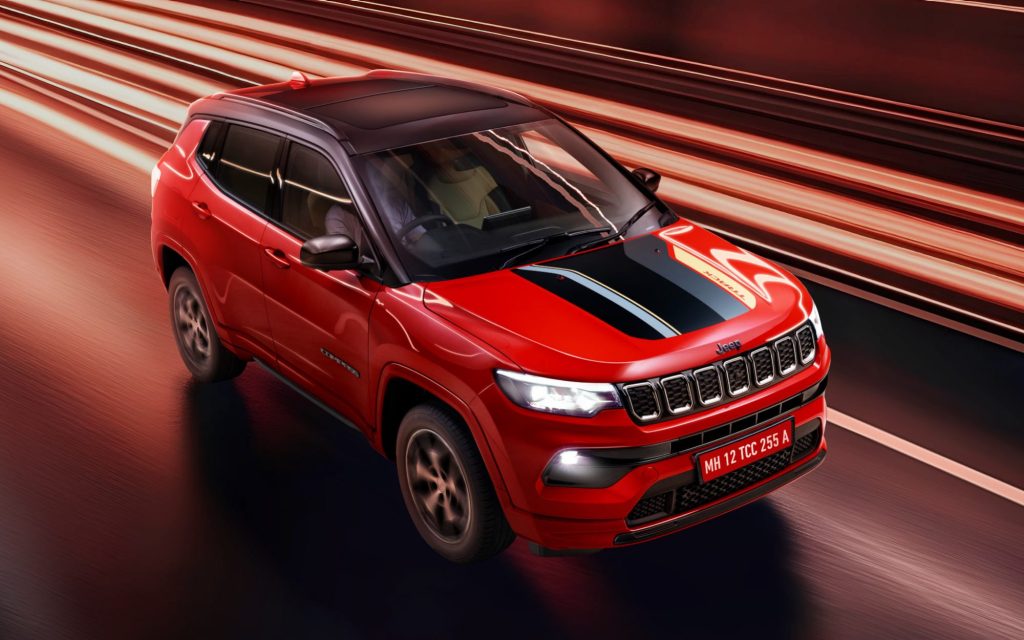 Dynamic front three-quarter view of the 2026 Jeep Compass in red with black hood accents, driving through illuminated motion blur at night.