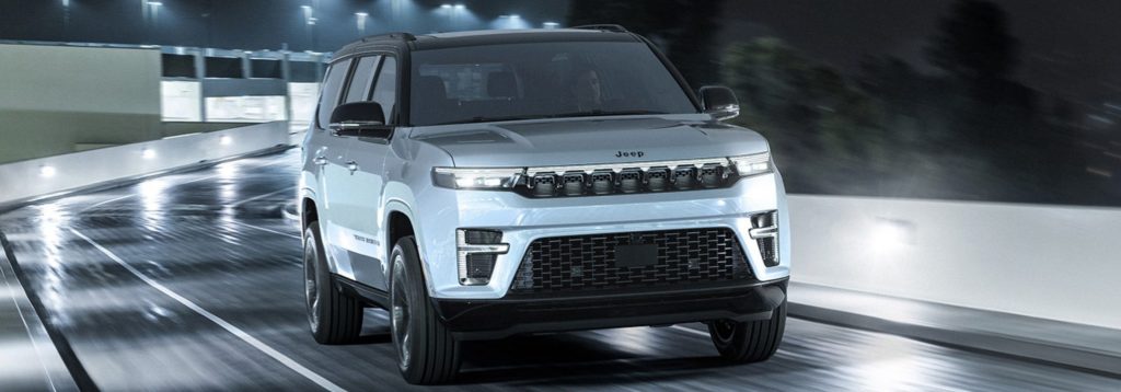 A white 2026 Jeep Grand Wagoneer drives at night on a wet, illuminated roadway, with reflections shimmering on the pavement.